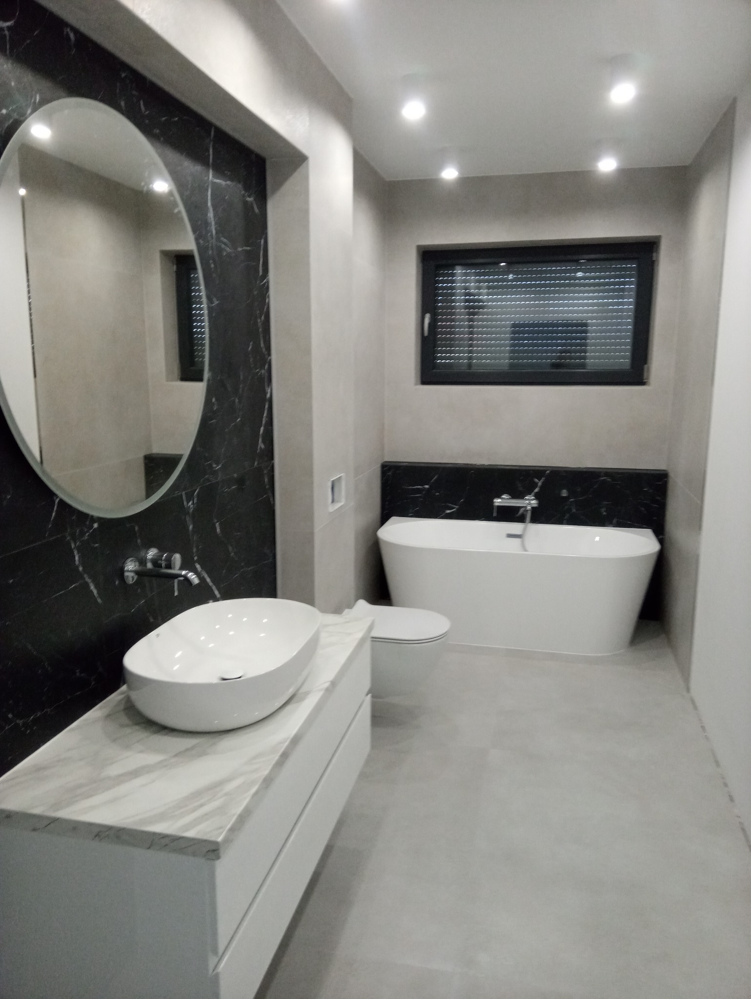 Nowoczesna łazienka z owalną umywalką nablatową na białej szafce z marmurowym blatem, owalne lustro na ścianie z czarnym marmurem, wolnostojąca wanna i podwieszana toaleta.