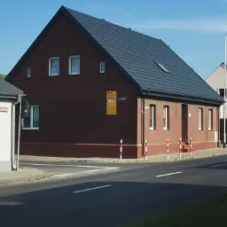 Dwukondygnacyjny dom z ciemną dachówką, widoczny od strony ulicy w słoneczny dzień. Na ścianie budynku tabliczka z adresem i ogłoszenie.