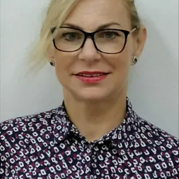 Portret uśmiechniętej blondynki w okularach i wzorzystej koszuli, na jasnym tle.