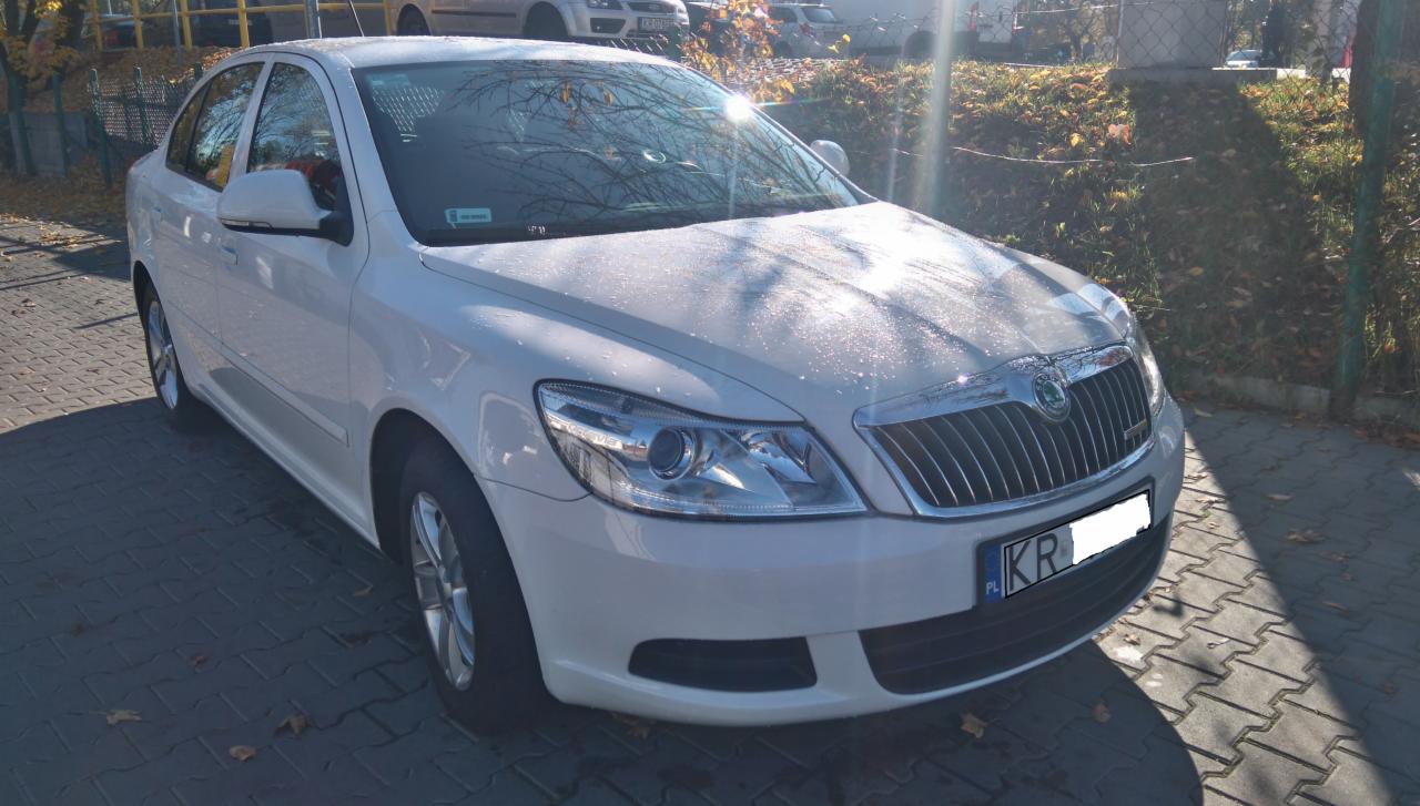 Biała Skoda Octavia z krakowską rejestracją KR, zaparkowana na brukowanym chodniku w słoneczny dzień. Widoczne jesienne liście i elementy zieleni w tle.