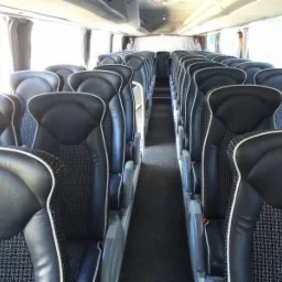 Wnętrze autobusu z rzędami czarnych foteli z jasnymi lamówkami i wzorzystymi wstawkami. Widok na przejście między siedzeniami, okna z zasłonami w tle.