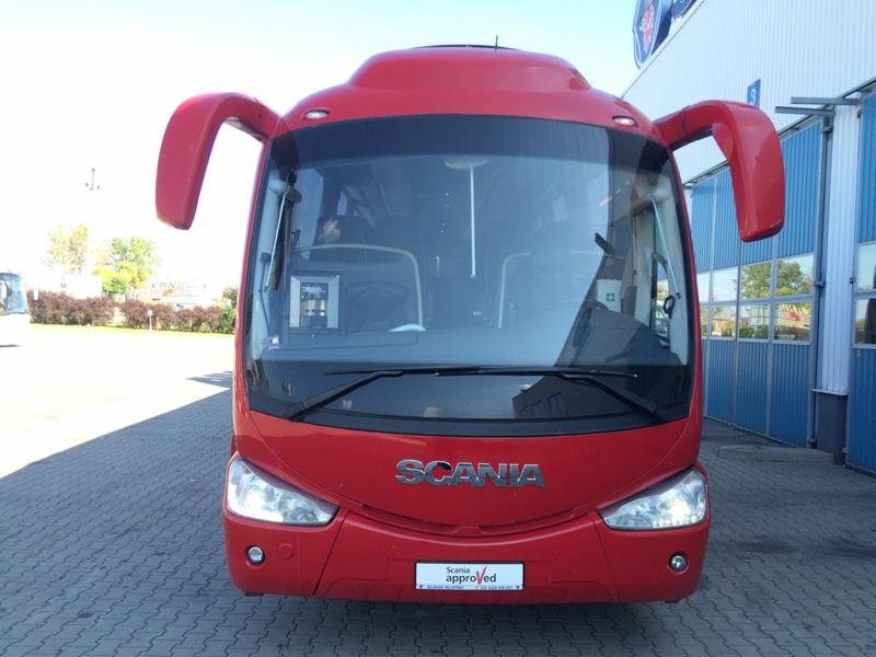 Czerwony autobus Scania z przodu, zaparkowany na zewnątrz, z widocznymi detalami karoserii i logo, na tle budynku z niebieskimi elementami.