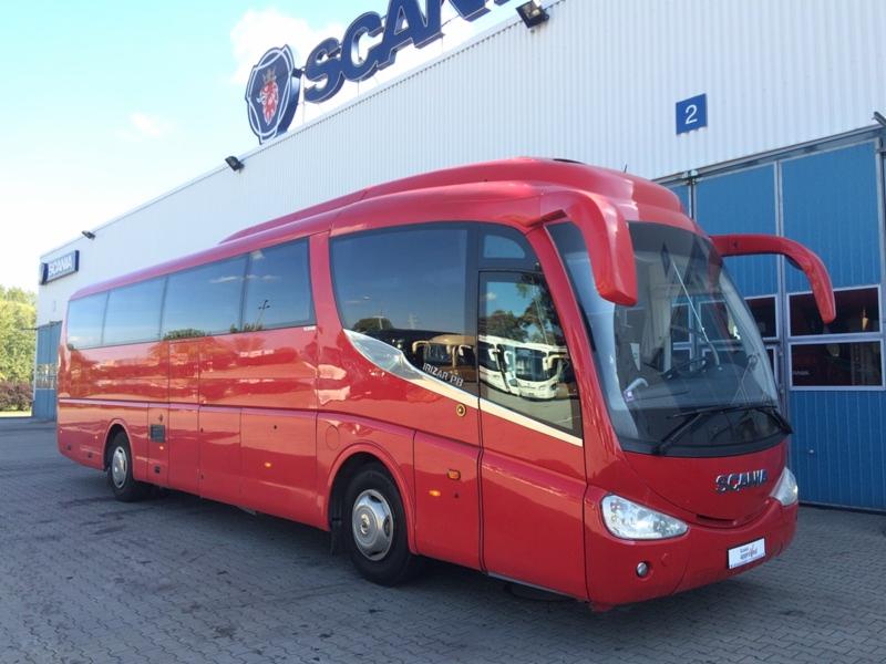 Czerwony autokar Scania Irizar na tle budynku z logo Scania i numerem 2. Widok z boku, oświetlenie dzienne, czyste szyby.