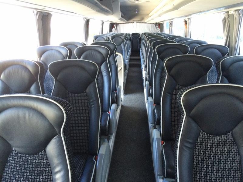 Wnętrze autobusu z rzędami czarnych foteli z jasnymi lamówkami i wzorzystymi wstawkami. Widok na przejście między siedzeniami, okna z zasłonami w tle.