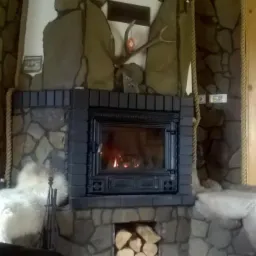 Rustykalny kominek z paleniskiem, obudowany kamieniem i wykończony drewnem w ciepłym odcieniu. Ozdobiony porożem jelenia i linami jutowymi, tworzy przytulny klimat.