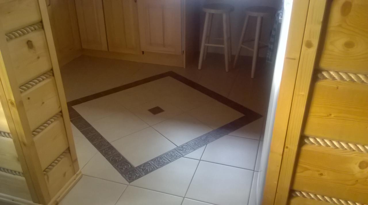 Wykończona podłoga w kuchni z jasnych płytek, obramowana ciemniejszym wzorem. Widoczne drewniane elementy wykończeniowe i stołki barowe w tle.