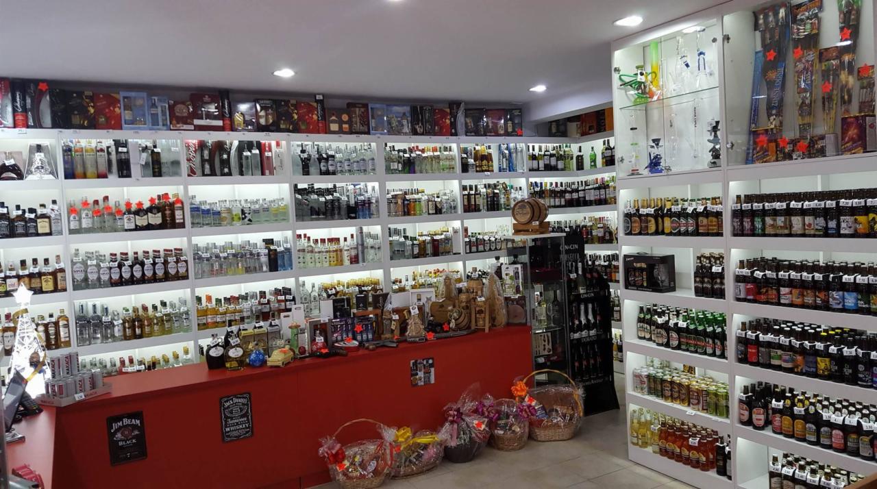 Wnętrze sklepu z alkoholem, wypełnione półkami z różnorodnymi butelkami i zestawami prezentowymi, z czerwonym blatem lady i koszami upominkowymi na pierwszym planie.
