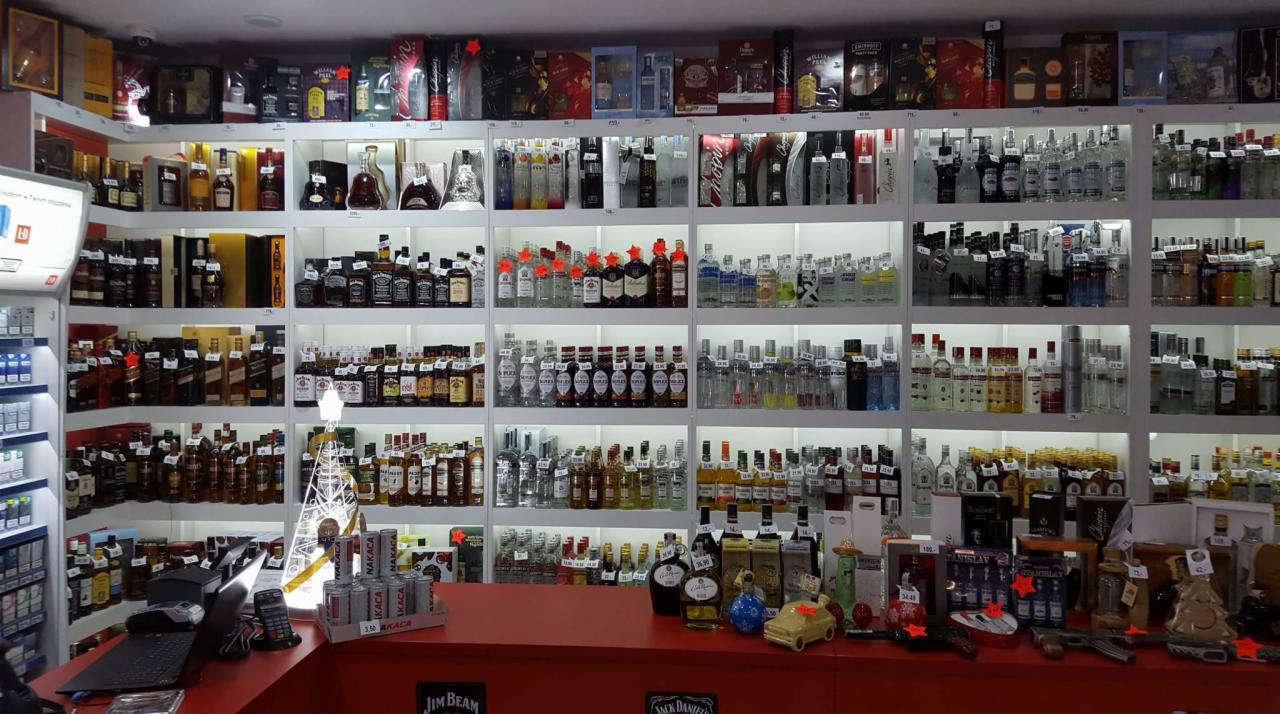 Półki sklepowe wypełnione różnorodnymi butelkami alkoholi, widoczne również elementy dekoracyjne na ladzie, takie jak ozdobne butelki i zabawkowy samochód.