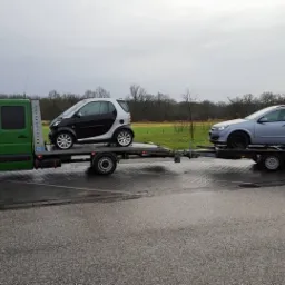 Zielona laweta z dwoma samochodami: Smart i Opel Astra kombi. Transport aut na mokrym asfalcie, pochmurne niebo w tle. Widok z boku, laweta z podwójną osią.