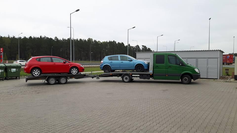 Zielona laweta z dwoma samochodami osobowymi: czerwonym i niebieskim. Laweta stoi na parkingu z kostki, w tle widoczne drzewa i budynki.