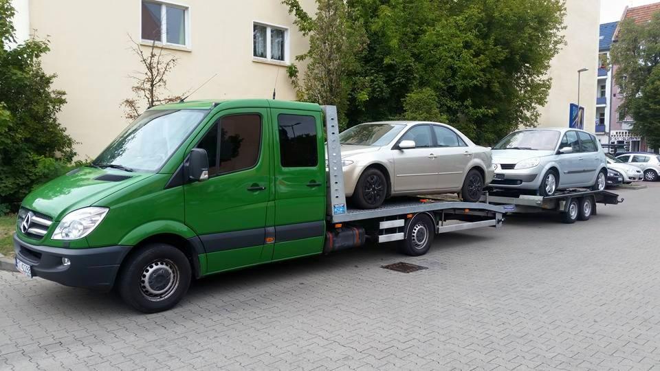 Zielona laweta Mercedes z dwoma samochodami (złoty sedan i srebrny hatchback) na przyczepie, zaparkowana na brukowanej ulicy w słoneczny dzień.