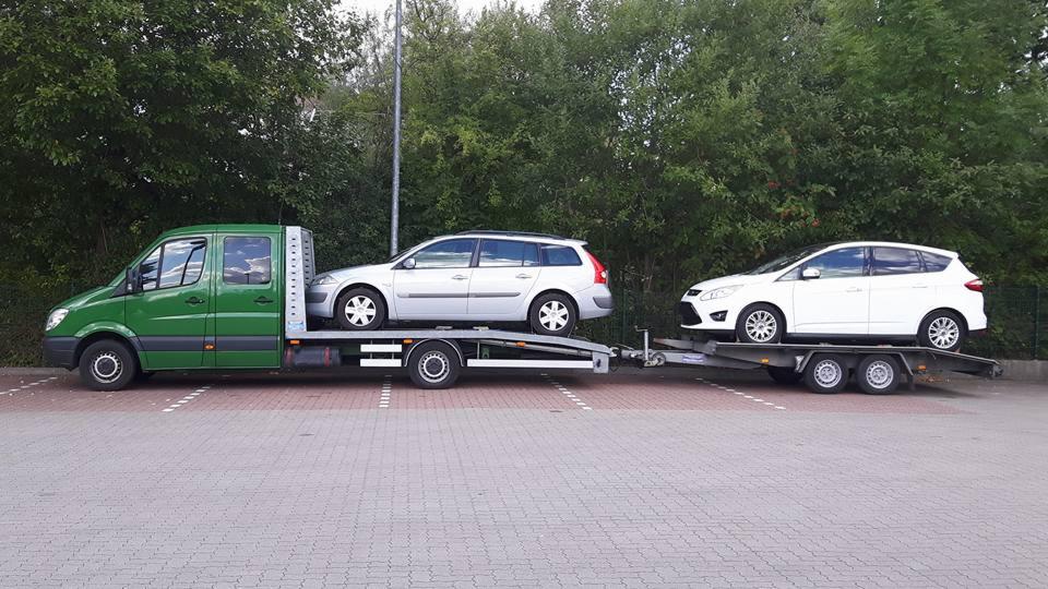 Zielona laweta z dwoma samochodami: srebrnym kombi i białym hatchbackiem, zaparkowana na parkingu z zielonymi drzewami w tle. Widok z boku, lekko z góry.