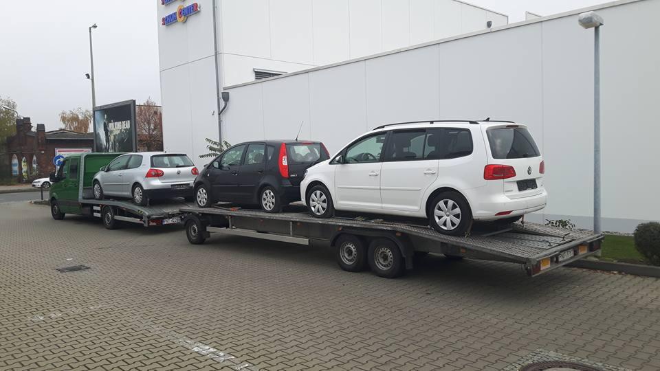 Laweta z trzema samochodami marki Volkswagen na przyczepie, zaparkowana na brukowanym placu z budynkiem w tle.