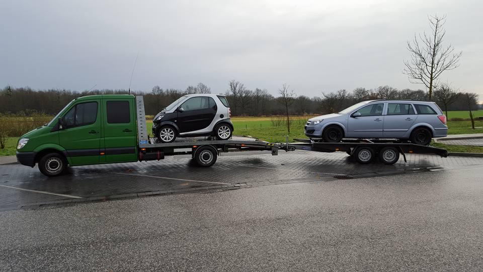 Zielona laweta z dwoma samochodami: Smart i Opel Astra kombi. Transport aut na mokrym asfalcie, pochmurne niebo w tle. Widok z boku, laweta z podwójną osią.