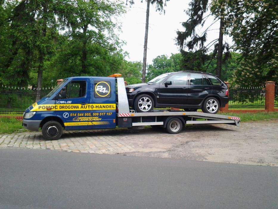 Laweta z czarnym SUV-em na platformie, zaparkowana na poboczu drogi, z widocznym logo i numerem telefonu pomocy drogowej na boku pojazdu.