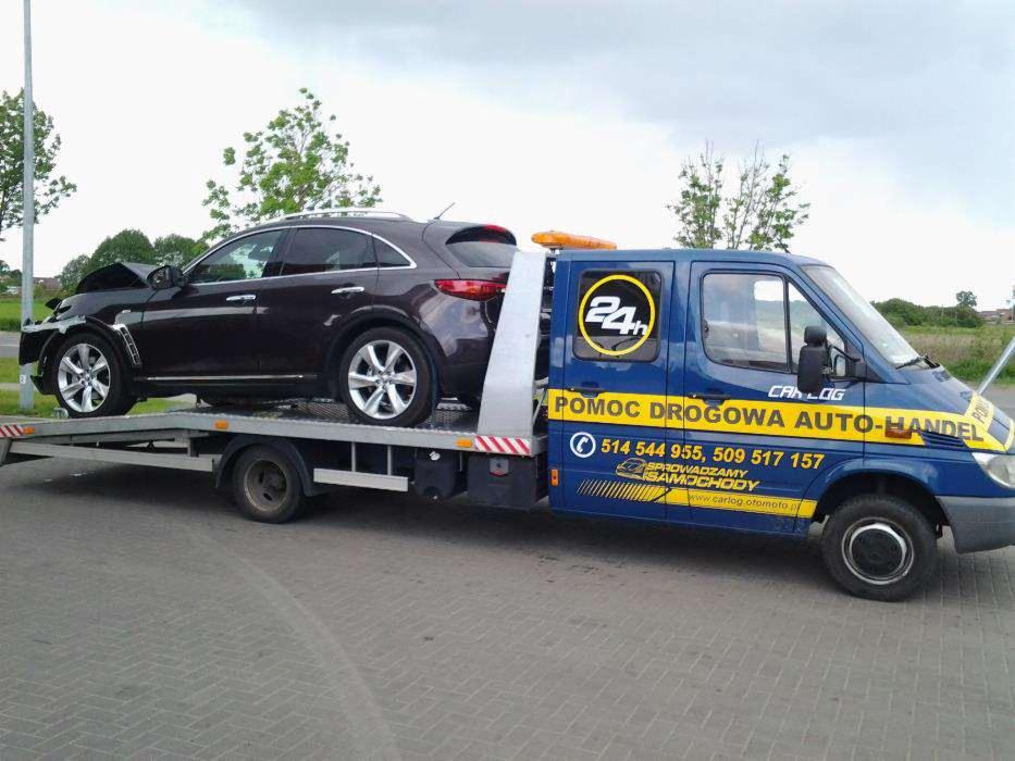 Laweta z uszkodzonym, brązowym SUV-em Infiniti FX na platformie. Pomoc drogowa Auto-Handel z numerami telefonów na niebieskiej kabinie. Widoczne uszkodzenia przodu pojazdu.