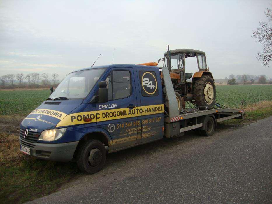 Laweta z ciągnikiem rolniczym na platformie, zaparkowana na poboczu drogi. Na boku pojazdu widoczne napisy 'Pomoc Drogowa Auto-Handel' i numery telefonów.