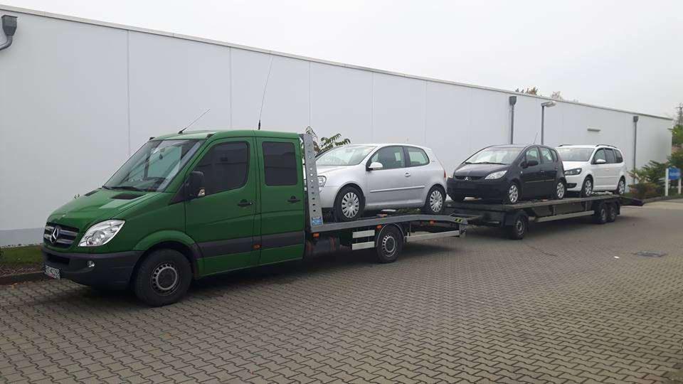 Zielona laweta Mercedes-Benz z trzema samochodami osobowymi na przyczepie, zaparkowana na brukowanym placu przed szarą halą z rynnami.