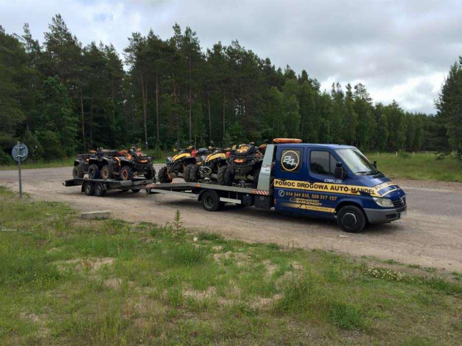 Niebieska laweta z przyczepą transportuje kilka quadów na tle leśnego krajobrazu.