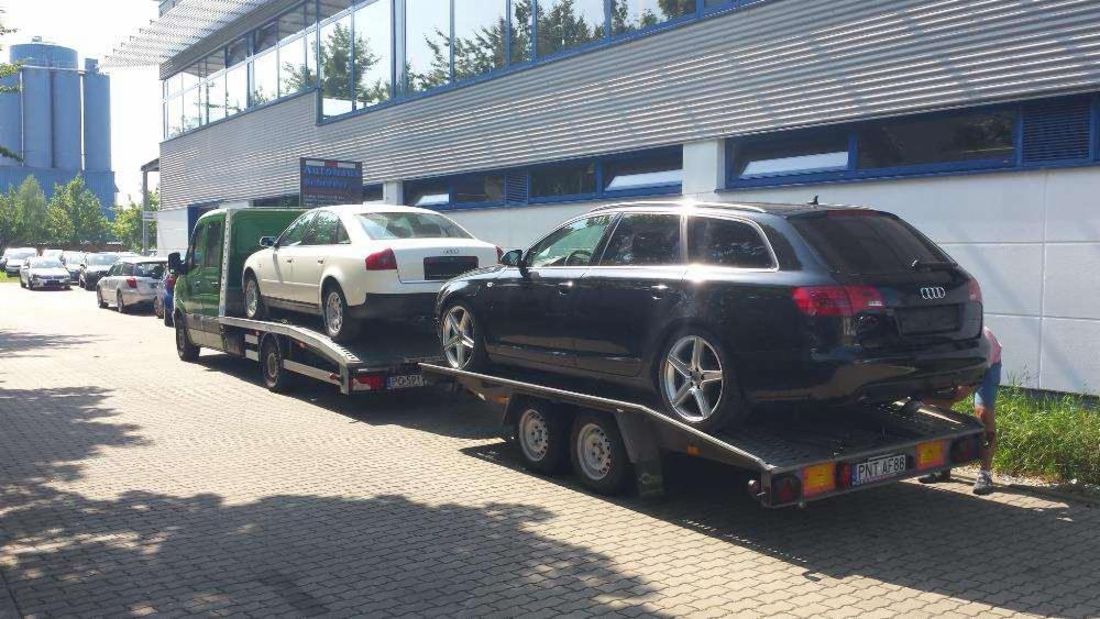 Laweta z dwoma samochodami Audi na przyczepie, zaparkowana przed budynkiem z szarymi żaluzjami. Widoczne tablice rejestracyjne.