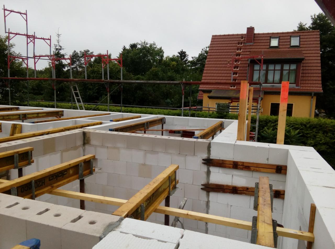 Wznoszenie ścian parteru z białych bloczków silikatowych, z drewnianymi stemplami i szalunkami, na tle żółtego domu z czerwoną dachówką i metalowymi rusztowaniami.