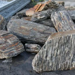 Dekoracyjne kamienie ogrodowe o unikalnej, warstwowej strukturze, ułożone na ziemi. W tle widoczny betonowy murek i drobny żwir.