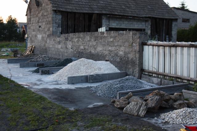 Przygotowane pryzmy kruszywa różnego rodzaju, oddzielone betonowymi obrzeżami na utwardzonym podłożu, w tle budynek gospodarczy i ogrodzenie.
