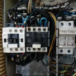 Rozdzielnia elektryczna z widocznymi stycznikami, bezpiecznikami i przewodami, wewnątrz skrzynki z metalową obudową, z widocznymi oznaczeniami producentów Siemens i Schneider Electric.