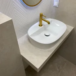 Biała, owalna umywalka nablatowa z baterią w kolorze złotym na blacie z płytek imitujących kamień, na ścianie dekoracyjne płytki z motywem jodełki i okrągłe lustro w złotej ramie, minimalistyczny...