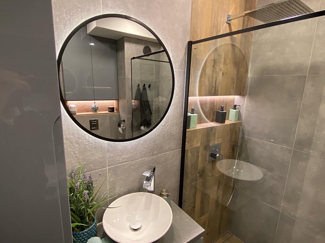Nowoczesna łazienka z okrągłym lustrem podświetlanym LED, białą umywalką nablatową, szarą armaturą, kabiną prysznicową typu walk-in i drewnianymi akcentami na ścianach.