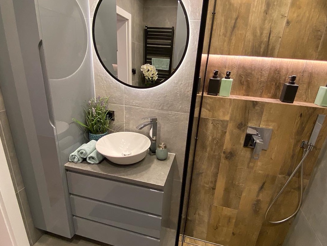 Nowoczesna łazienka z okrągłym lustrem podświetlanym LED, umywalką nablatową, szarą szafką z szufladami, drewnopodobnymi płytkami w strefie prysznica i dekoracyjnymi akcesoriami.
