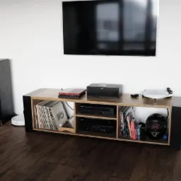 Nowoczesna szafka RTV z drewna i czarnych elementów, wypełniona płytami winylowymi, sprzętem audio i kaskiem motocyklowym, pod telewizorem na białej ścianie.