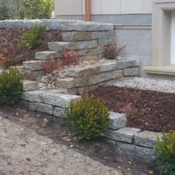 Granitowa kaskada w ogrodzie: kamienne murki tarasowe obsadzone zielenią, z korą i białym żwirem, tworzą harmonijną kompozycję przy budynku z oknem.