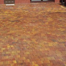 Sw-Bud sp. z.o. o - Umyta, nowa, wielobarwna kostka brukowa w odcieniach brązu i żółci, ułożona na podjeździe przed budynkiem z cegły. Osoba z wężem ogrodowym w lewym górnym rogu.