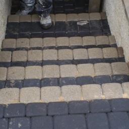 Sw-Bud sp. z.o. o - Ułożona kostka brukowa w dwóch kolorach na schodach wejściowych, obrzeża z kamiennych bloków, Radom. Widoczne buty osoby układającej kostkę.