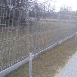 Sw-Bud sp. z.o. o - Nowoczesne ogrodzenie panelowe z podmurówką, widok z boku. Szara metalowa siatka, betonowa podmurówka, trawnik i kostka brukowa. W tle budynek i drzewa.