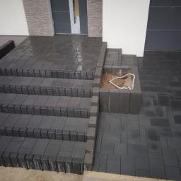 Sw-Bud sp. z.o. o - Schody wejściowe wyłożone ciemną kostką brukową, z donicą po prawej stronie, widoczne przewody w donicy, mokra nawierzchnia, nowoczesny design.