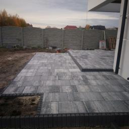 Sw-Bud sp. z.o. o - Szara kostka brukowa ułożona na tarasie, z widocznym obrzeżem z ciemniejszych kostek, częściowo jeszcze w trakcie prac, z ziemią widoczną w miejscach do uzupełnienia, w tle betonowe ogrodzenie...