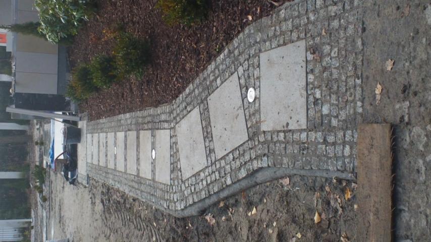 Ścieżka z kostki granitowej i płyt betonowych, oświetlona punktowymi lampami, prowadząca przez ogród. Widoczne elementy krajobrazu i narzędzia brukarskie.