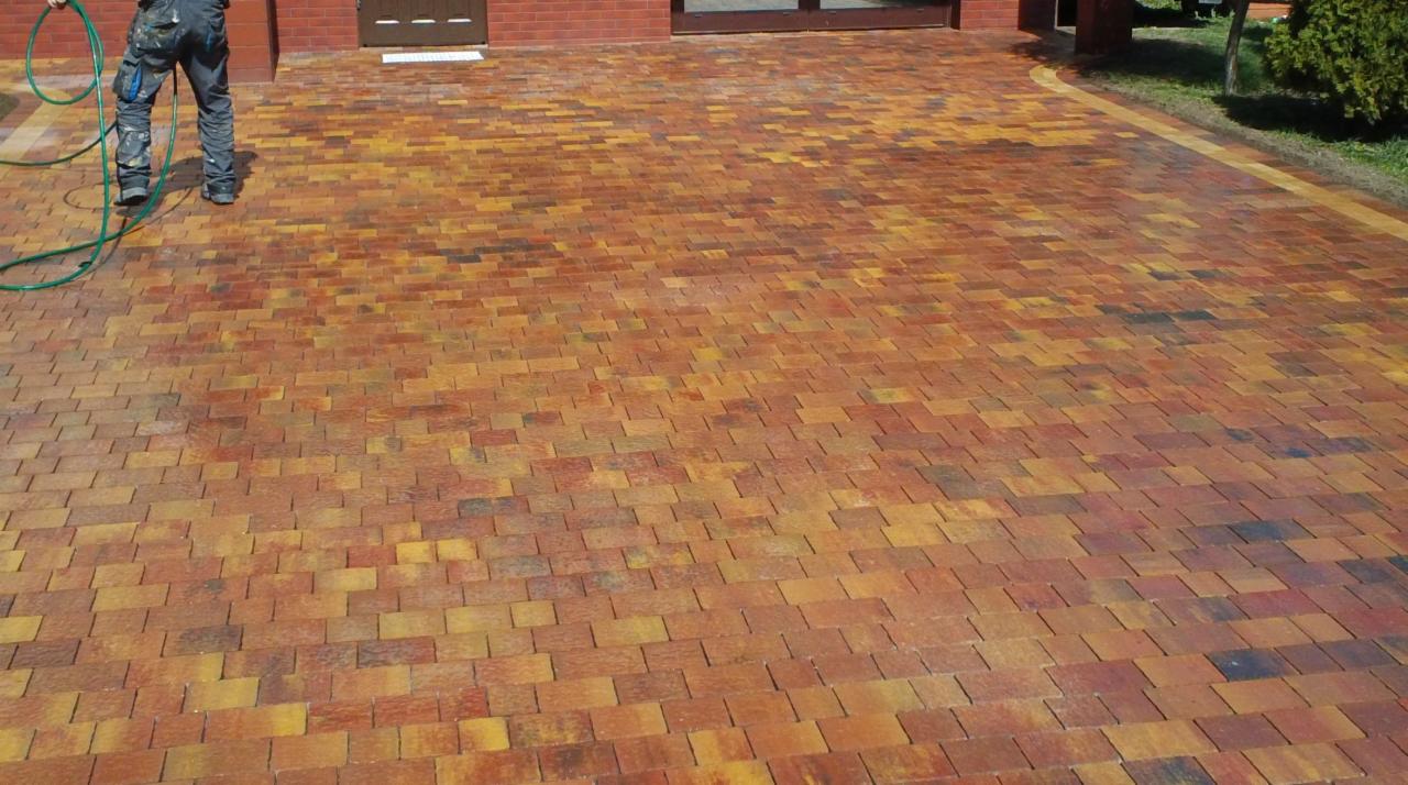 Umyta, nowa, wielobarwna kostka brukowa w odcieniach brązu i żółci, ułożona na podjeździe przed budynkiem z cegły. Osoba z wężem ogrodowym w lewym górnym rogu.