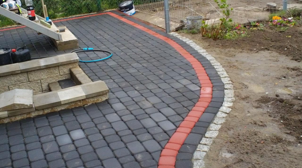 Ułożona kostka brukowa w kolorze szaro-czarnym z czerwonym obramowaniem. Widoczne schody z kostki i fragment trawnika z kwiatami. W tle siatka ogrodzeniowa.