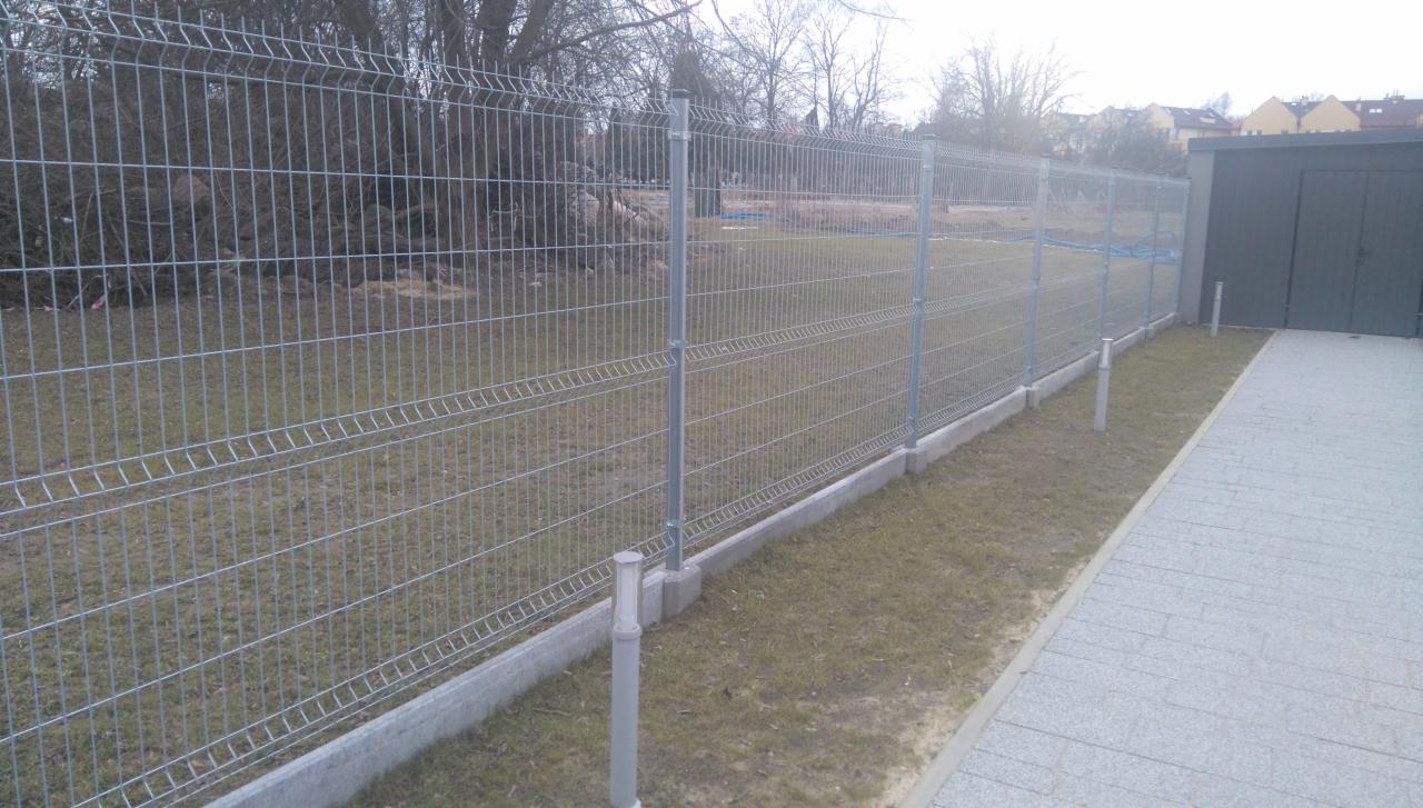 Nowoczesne ogrodzenie panelowe z podmurówką, widok z boku. Szara metalowa siatka, betonowa podmurówka, trawnik i kostka brukowa. W tle budynek i drzewa.