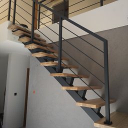 Kowalstwo Kubala - Nowoczesne schody z drewnianymi stopniami i czarną metalową konstrukcją oraz minimalistyczną balustradą, widoczne na tle szarej ściany z fakturą i elementami drewnianymi na suficie.
