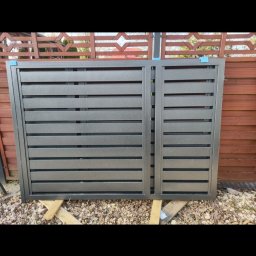 Kowalstwo Kubala - Fragment szarej, metalowej bramy wjazdowej z poziomymi panelami, oparty na drewnianych belkach na zewnątrz, z widocznym drewnianym ogrodzeniem w tle.