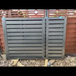 Fragment szarej, metalowej bramy wjazdowej z poziomymi panelami, oparty na drewnianych belkach na zewnątrz, z widocznym drewnianym ogrodzeniem w tle.