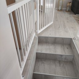 Kowalstwo Kubala - Biała barierka zabezpieczająca schody wewnętrzne z drewnopodobnymi stopniami, widoczna z perspektywy góry, z częściowo otwartymi drzwiczkami. W tle elementy wystroju wnętrza.