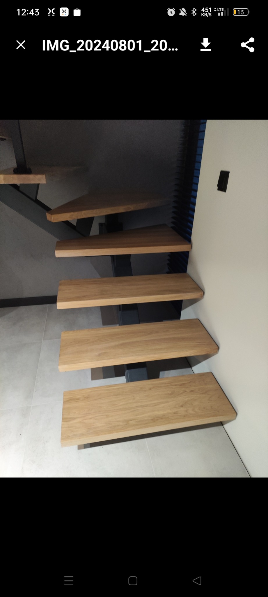 Nowoczesne schody z drewnianymi stopniami i metalową konstrukcją nośną, widok z dołu, jasne płytki podłogowe i biała ściana w tle.