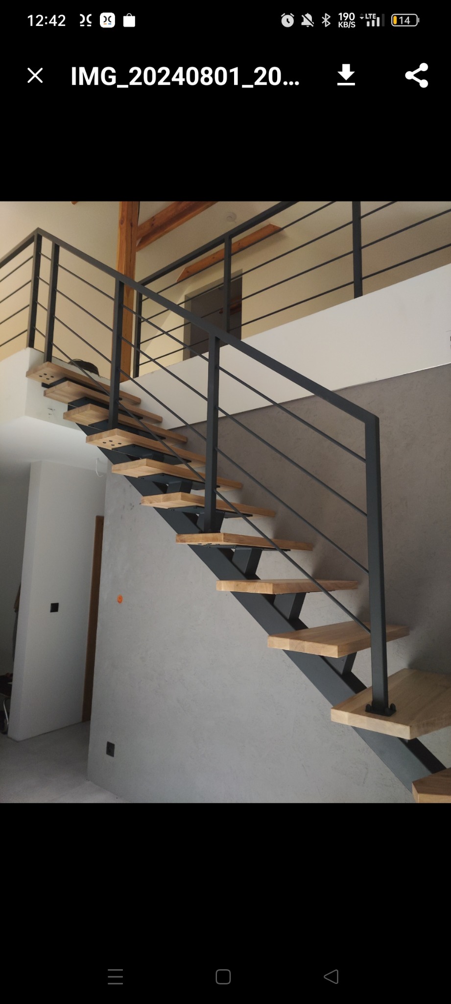 Nowoczesne schody z drewnianymi stopniami i czarną metalową konstrukcją oraz minimalistyczną balustradą, widoczne na tle szarej ściany z fakturą i elementami drewnianymi na suficie.