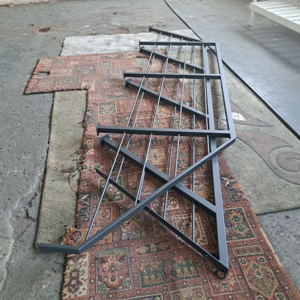 Element metalowej balustrady schodowej w kolorze szarym, leżący na dywanie w warsztacie przed montażem.