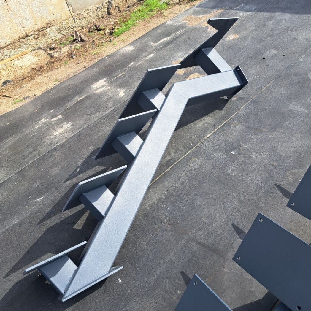 Surowe, metalowe schody modułowe w kolorze szarym, przygotowane do montażu na zewnątrz, widoczne mocowania, surowy beton w tle.